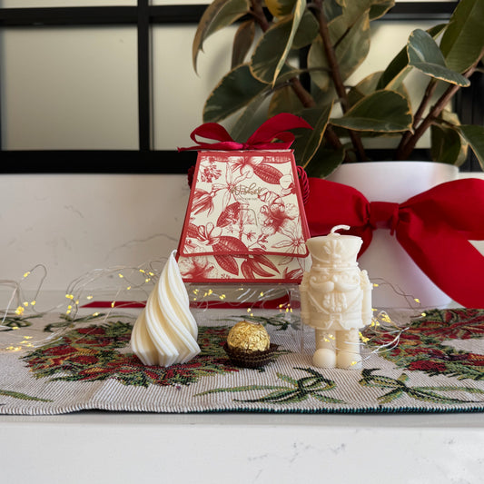 A styled holiday tabletop scene with a red and white patterned gift box tied with a red ribbon, a cream astronaut candle, a cream spiral candle, a small gold ball, and a floral runner, set against a white wall with a window frame and potted green plants.
