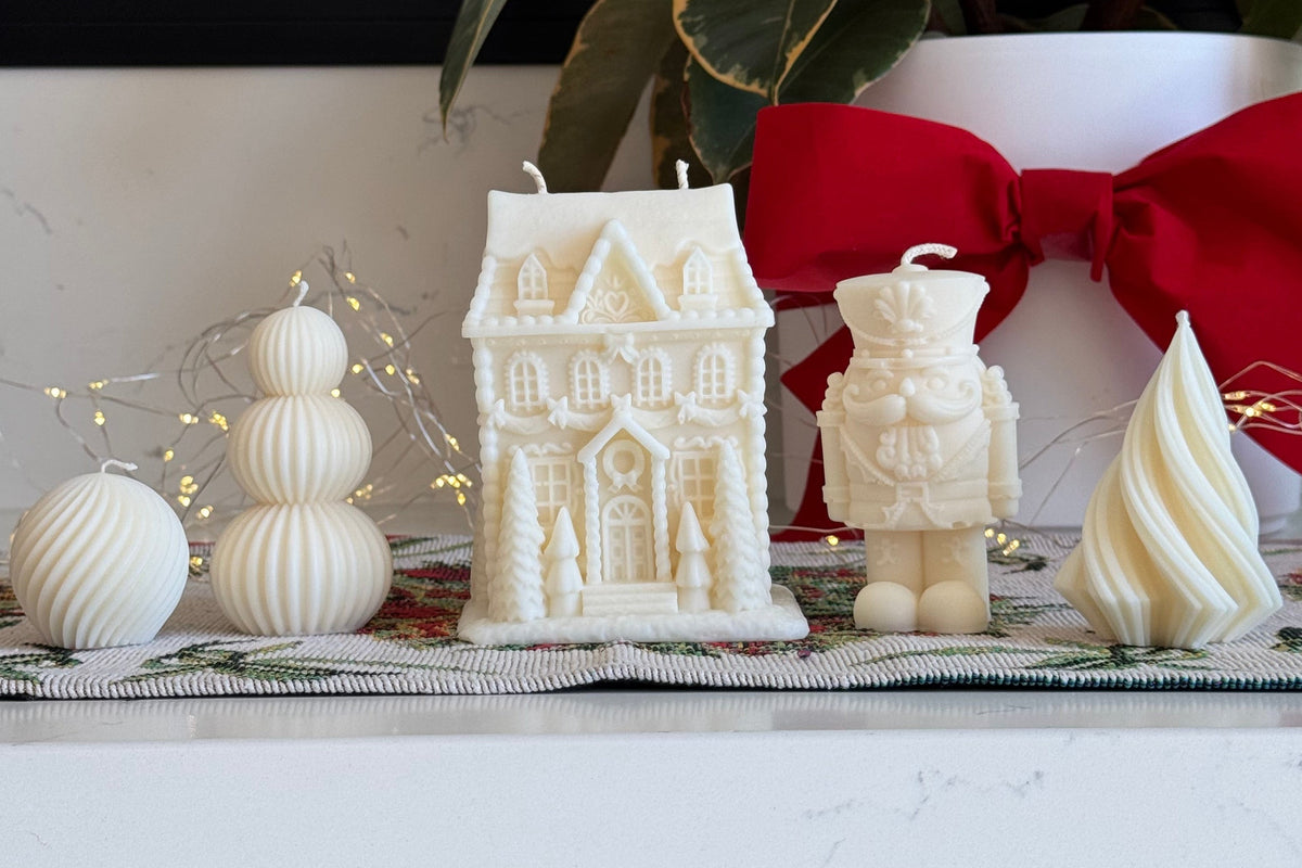 A collection of white holiday-themed decorative candles displayed on a white marble countertop, including a gingerbread house candle, a nutcracker candle, a spiral tree candle, and stacked sphere candles, with a red bow and green foliage in the background.
