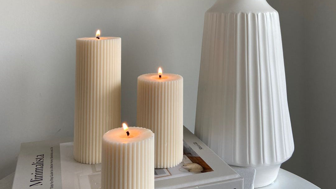 Three ribbed candles and a white vase with dried flowers on a surface with a neutral background.