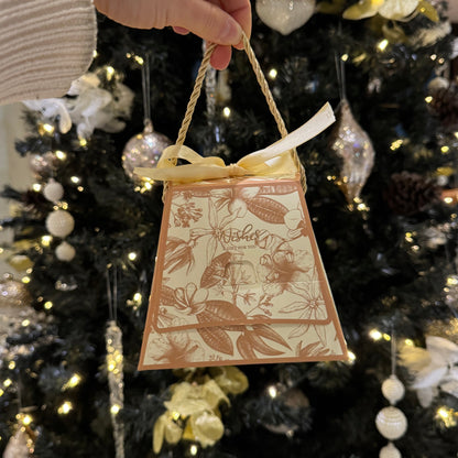 A hand wearing a white ribbed sweater holds a small decorative gift bag in front of a decorated Christmas tree. The bag is cream-colored with a brown botanical illustration and a yellow ribbon tied in a bow at the top.