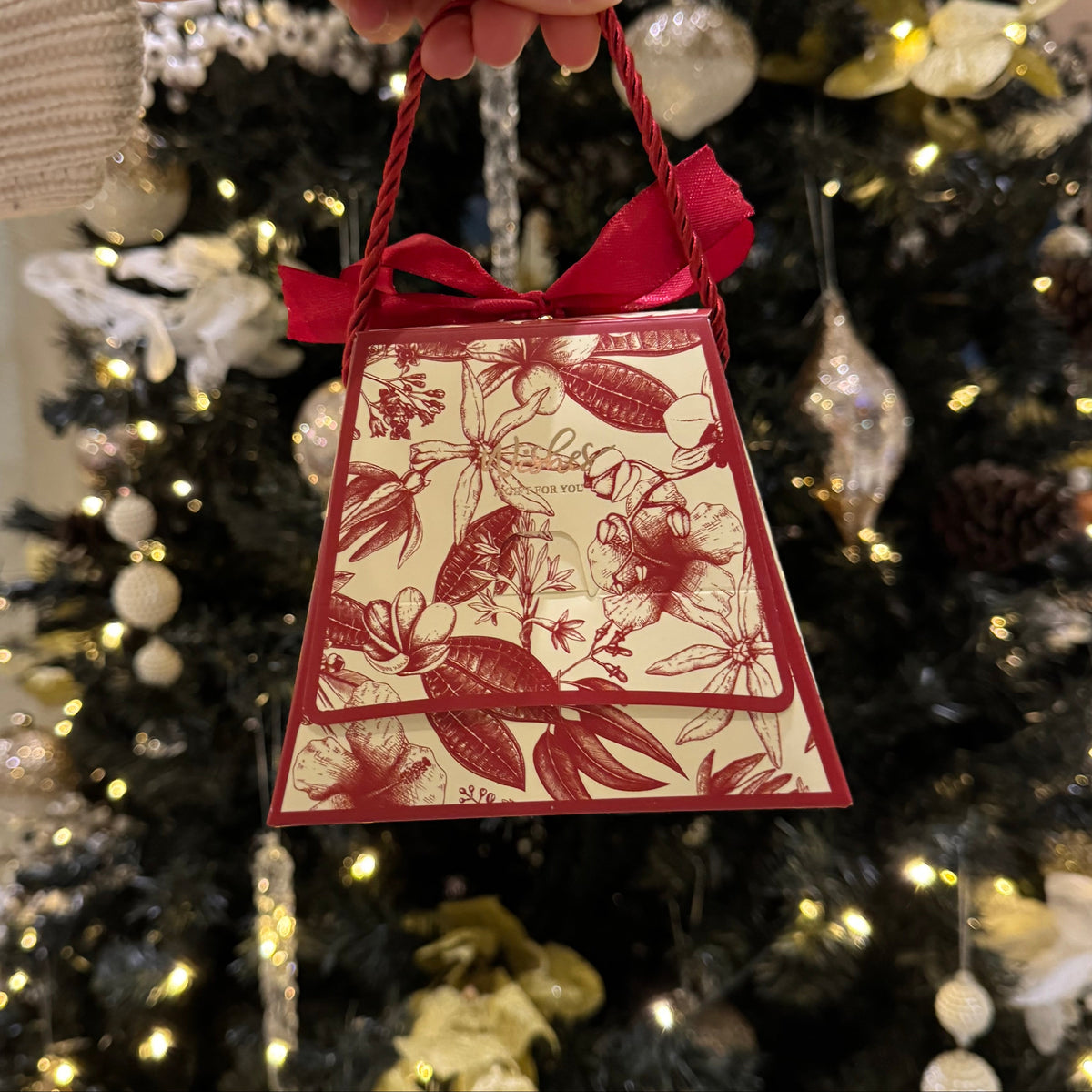 A hand holding a small decorative gift bag with red ribbon handles, featuring a red floral toile pattern on a cream background, set against a blurred Christmas tree with lights and ornaments.