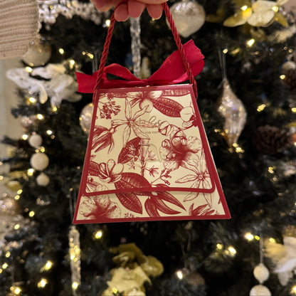 A hand holding a small decorative gift bag with red ribbon handles, featuring a red floral toile pattern on a cream background, set against a blurred Christmas tree with lights and ornaments.