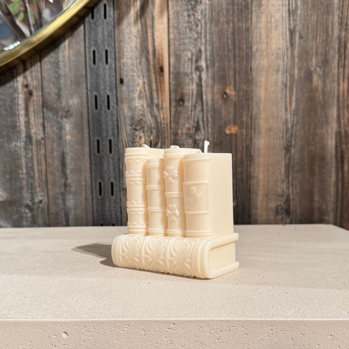 A set of four cream-colored book-shaped candles arranged on a light stone surface in front of a rustic wooden wall.