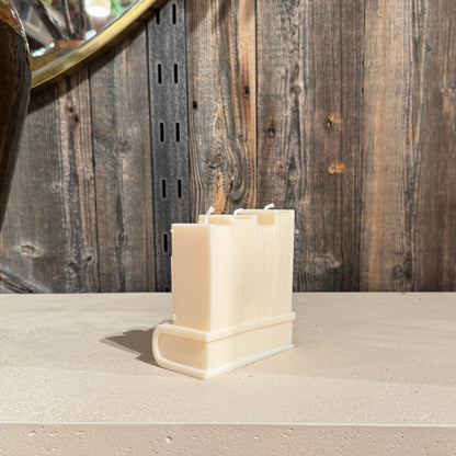 A cream-colored book shaped candle, displayed on a light stone countertop against a rustic wood-paneled wall.