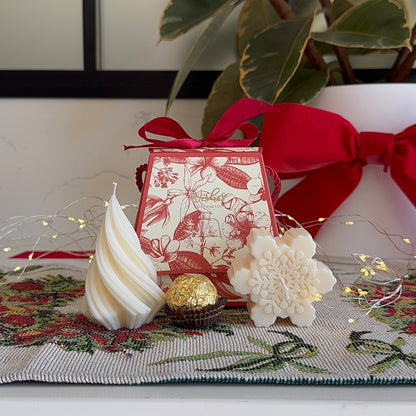 A festive holiday-themed product display featuring a small red and white patterned gift box tied with a red ribbon, alongside a white swirled meringue, a white flower-shaped confection, and a Ferrero Rocher chocolate, arranged on a patterned runner with string lights.