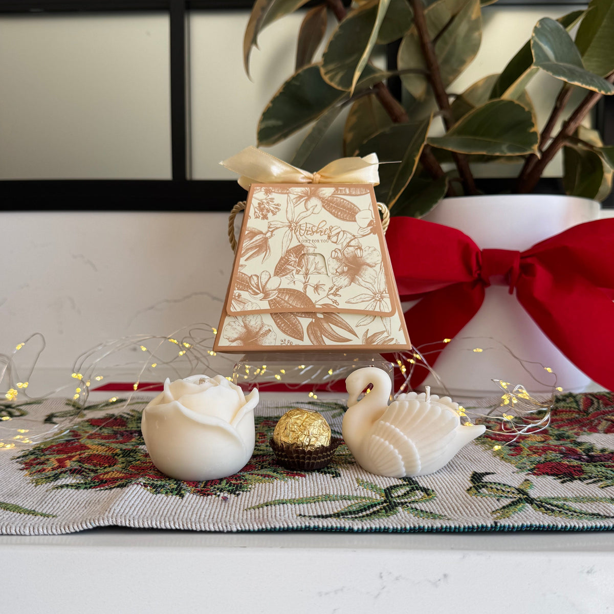 A decorative cream-colored gift box with a floral pattern and ribbon sits on a holiday-themed table runner alongside a white rose, a swan figurine, a small gold ball, and a large red bow on a white planter in the background.