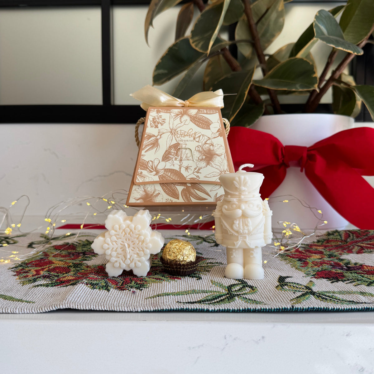 A styled tabletop display featuring a cream-colored astronaut candle, a small cream patterned gift box with a gold ribbon, a white flower, a gold ball, and a white pot with a large red bow in the background.