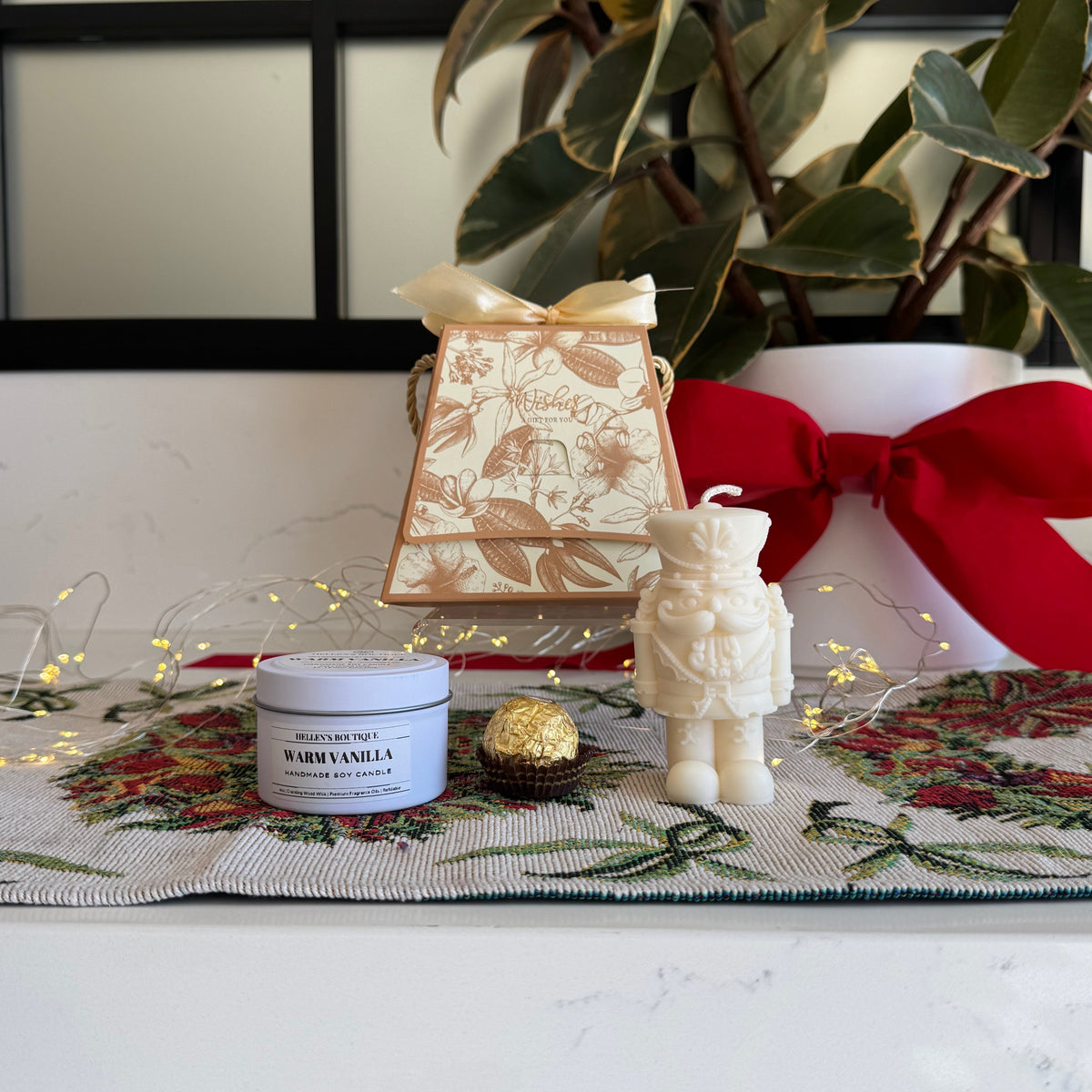 A styled holiday gift set on a white marble countertop, featuring a white jar of 'Warm Vanilla' scented candle, a cream nutcracker-shaped candle, a small gold foil-wrapped chocolate, and a cream gift bag tied with a red ribbon, set against a background of a potted plant.