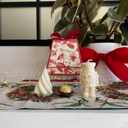 A styled holiday tabletop scene with a red and white patterned gift box tied with a red ribbon, a cream astronaut candle, a cream spiral candle, a small gold ball, and a floral runner, set against a white wall with a window frame and potted green plants.