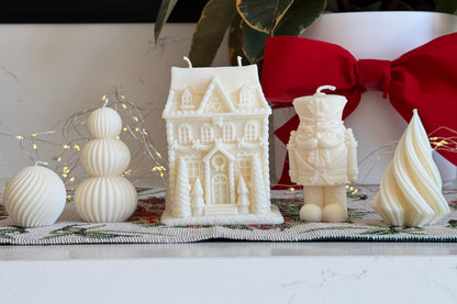 A collection of white holiday-themed decorative candles displayed on a white marble countertop, including a gingerbread house candle, a nutcracker candle, a spiral tree candle, and stacked sphere candles, with a red bow and green foliage in the background.