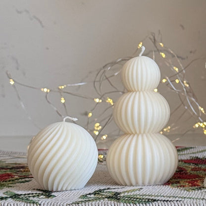 A collection of white holiday-themed candles displayed on a white marble countertop, including a gingerbread house candle, a nutcracker candle, a spiral tree candle, and stacked sphere candles, with a red bow and green foliage in the background.