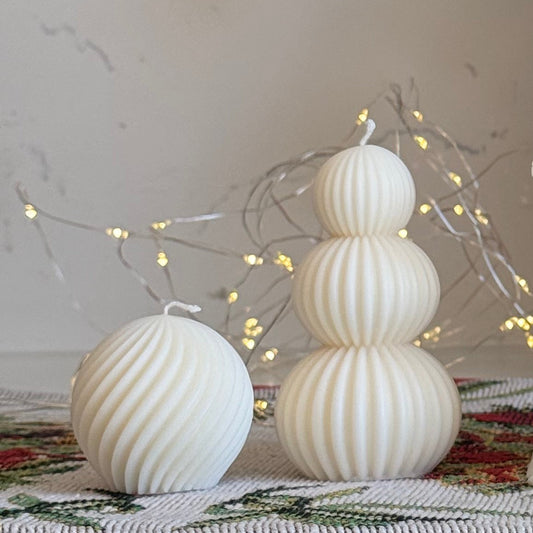 A collection of white holiday-themed candles displayed on a white marble countertop, including a gingerbread house candle, a nutcracker candle, a spiral tree candle, and stacked sphere candles, with a red bow and green foliage in the background.