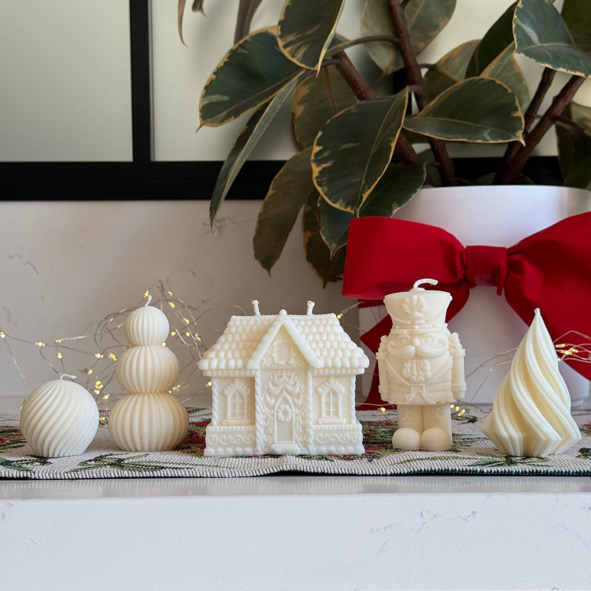 A collection of white holiday-themed candles displayed on a white countertop, including a gingerbread house-shaped candle, a nutcracker figure candle, a spiral tree candle, and stacked snowball candles, with a red bow and a potted variegated plant in the background.