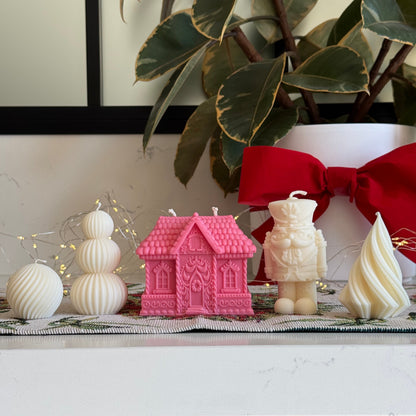 A styled holiday display featuring a pink gingerbread house-shaped candle, a cream nutcracker figurine, a cream swirled candle, and white spherical ornaments on a tabletop with a green plant and red bow in the background.