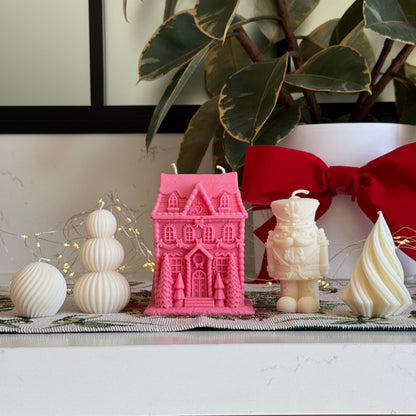 A collection of decorative candles displayed on a white marble surface, including a pink gingerbread house-shaped candle, a cream nutcracker figure candle, a cream spiral candle, and white spherical ornaments, with a large red bow and green houseplant in the background.