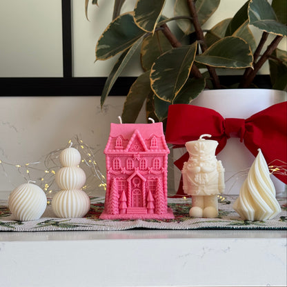 A styled holiday candle display on a white surface, featuring a prominent pink gingerbread house-shaped candle, a cream nutcracker figurine, a cream swirled candle, and white ribbed ornaments, with a red bow and green plant leaves in the background.