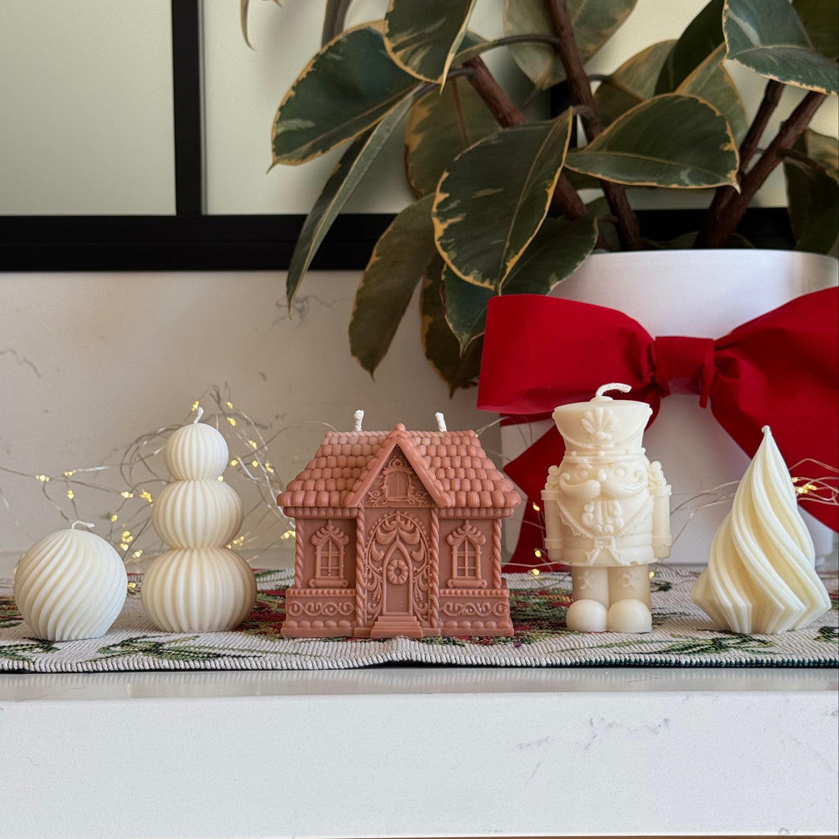A collection of Christmas-themed candles displayed on a white marble countertop, including a gingerbread house candle, a nutcracker candle, a snowman candle, and a white swan candle, with a potted variegated plant and a red bow in the background.