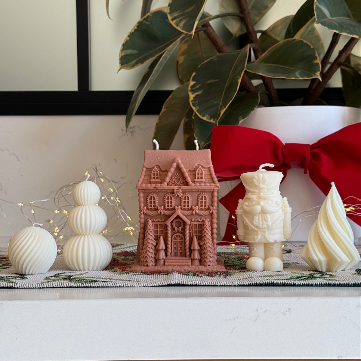 A styled holiday tabletop display featuring a light brown gingerbread house candle, a cream nutcracker candle, a cream swirled candle, and white spherical ornaments, with a large red bow and green variegated plant leaves in the background.