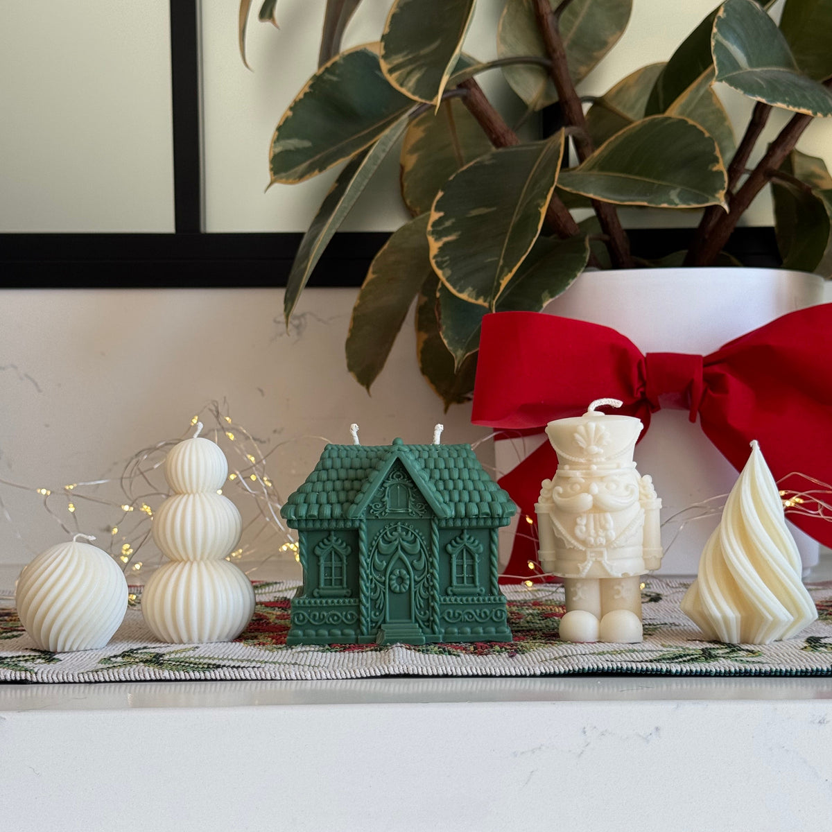 A set of Christmas-themed soy candles displayed on a white mantel. The set includes a green gingerbread house-shaped candle, a cream-colored nutcracker candle, a cream Christmas tree candle, and three cream ribbed snowball ornaments.