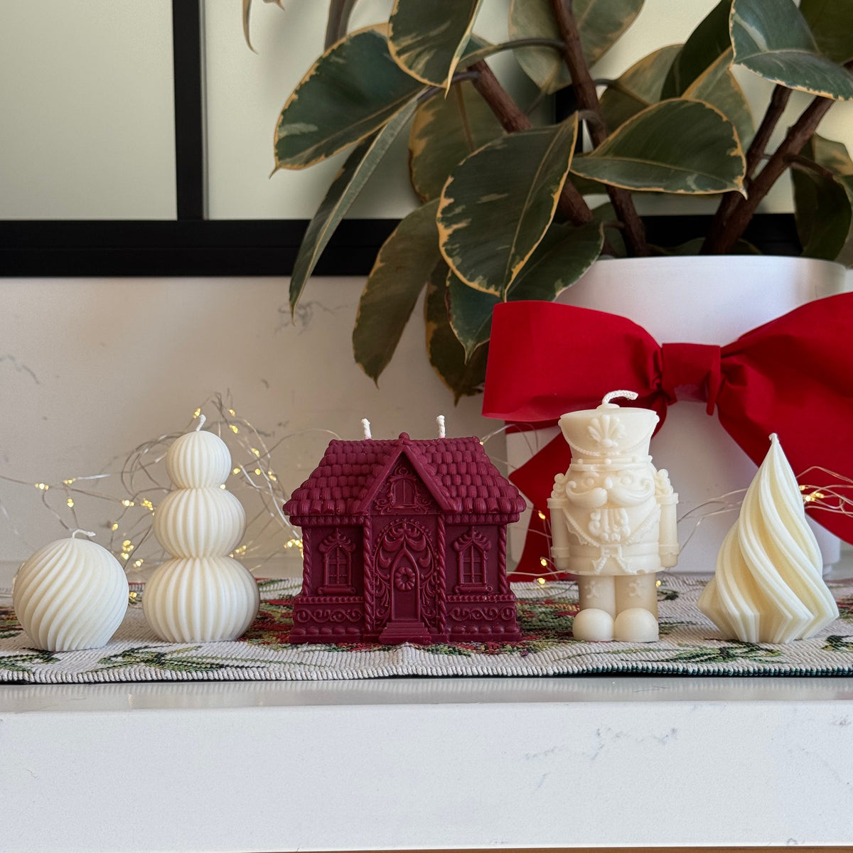 A collection of Christmas-themed candles displayed on a white marble countertop, including a burgundy red gingerbread house-shaped candle, a cream soldier candle, a cream meringue candle, and white spherical ornaments, with a potted variegated plant and a red bow in the background.