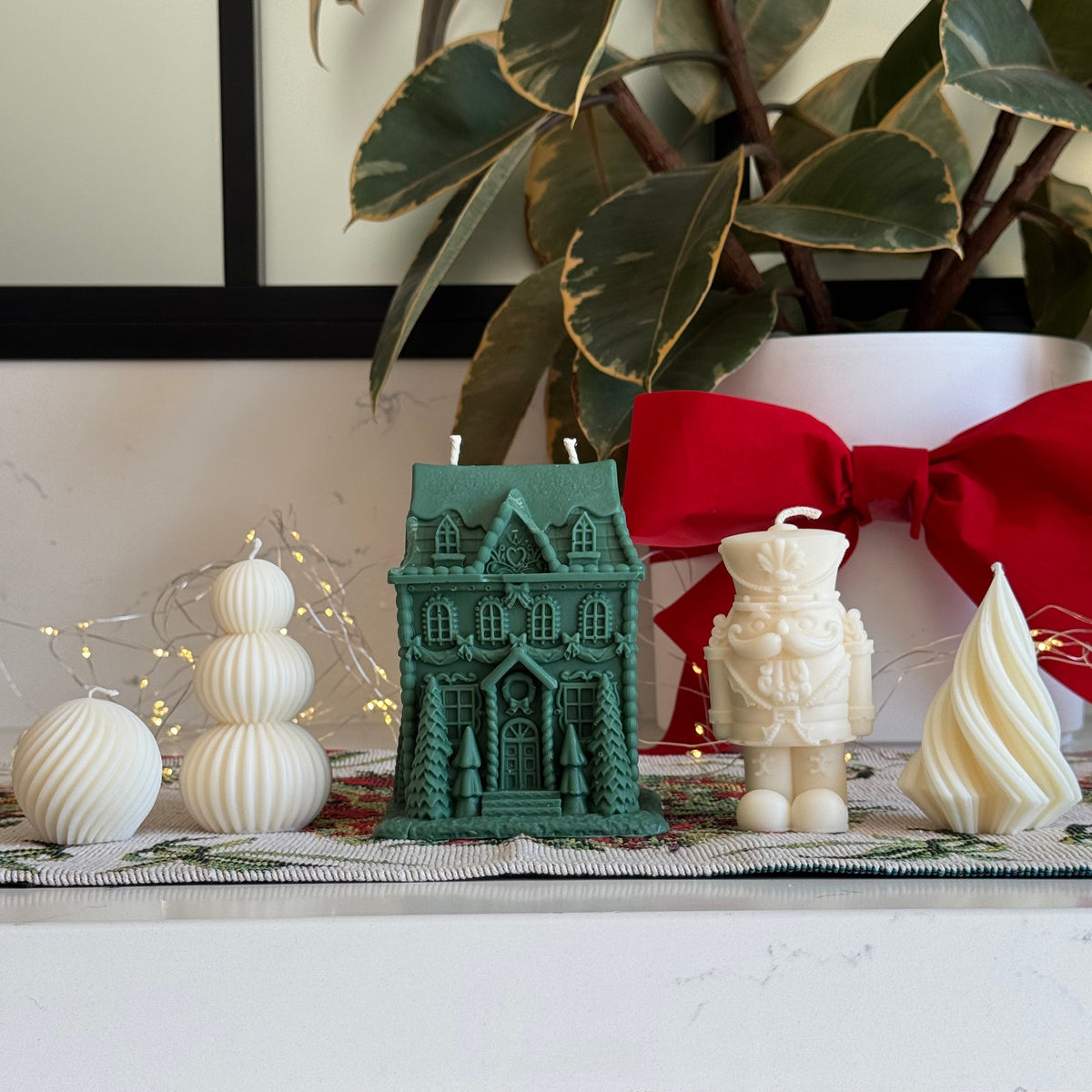A styled holiday tabletop display featuring a green gingerbread house-shaped candle, an off-white nutcracker figurine, a cream swirled candle, white spherical ornaments, and a large red bow in the background, with a potted variegated plant and a marble countertop.