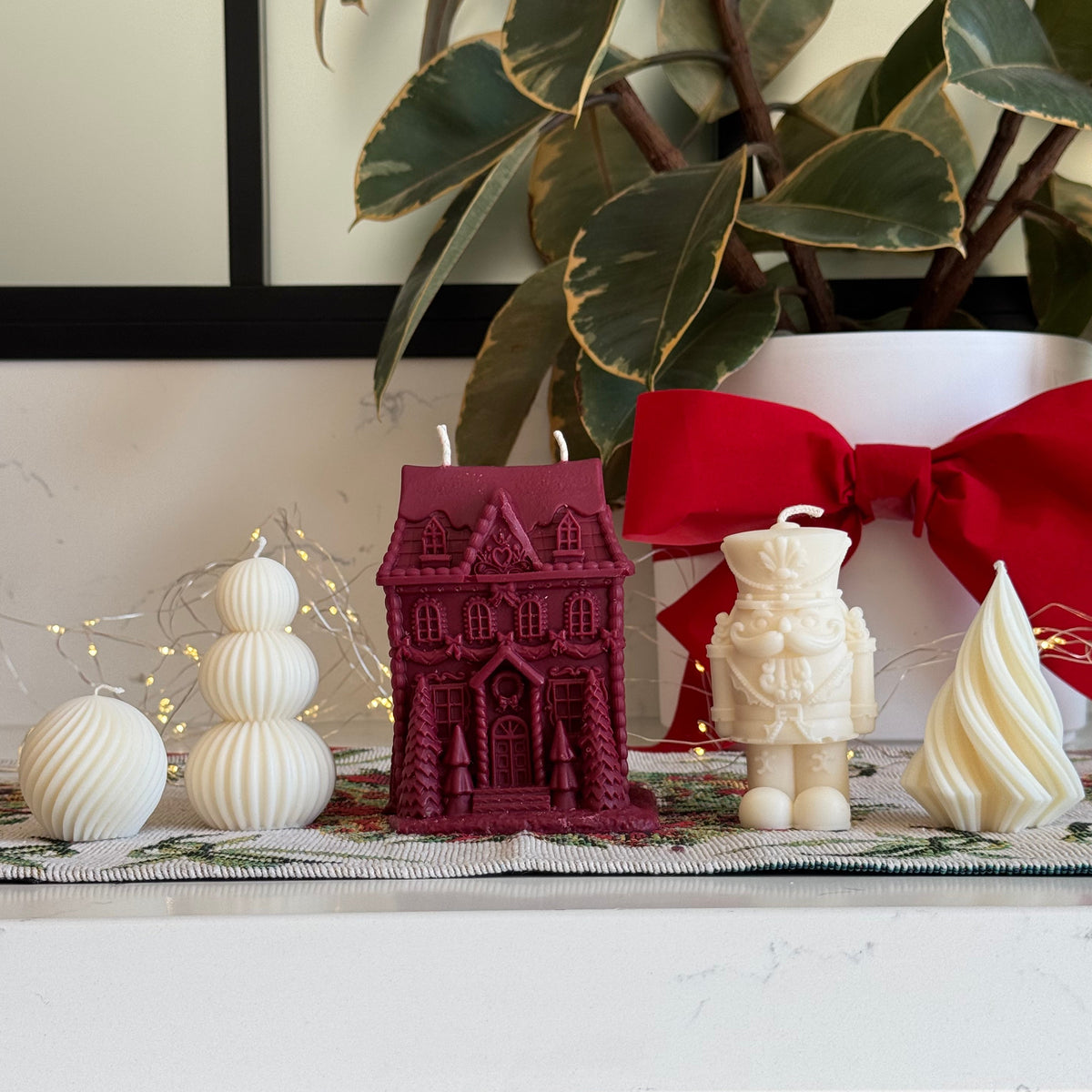 A collection of decorative Christmas-themed candles displayed on a white marble countertop. The set includes a tall, burgundy gingerbread house-shaped candle, a cream-colored nutcracker candle, a cream spiral tree candle, white ribbed spherical ornaments, and a large cream candle tied with a red ribbon.