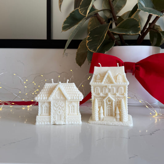 Two white holiday gingerbread house-shaped candles displayed on a white marble surface, with a red ribbon bow and a potted rubber plant in the background.