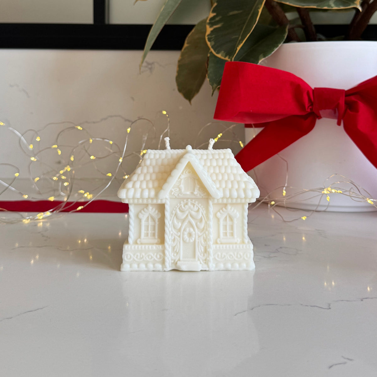 A white, gingerbread house-shaped candle sits on a light marble surface in front of a potted plant with variegated green leaves and a large red ribbon. Warm fairy lights are draped around the base.