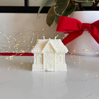 A white, gingerbread house-shaped candle sits on a light marble surface in front of a potted plant with variegated green leaves and a large red ribbon. Warm fairy lights are draped around the base.