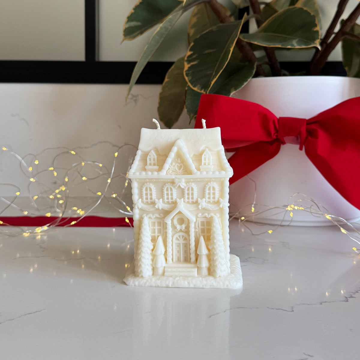 A large white gingerbread house-shaped candle sits on a light marble surface, styled with warm fairy lights, a potted plant with a red ribbon, and a white vase.