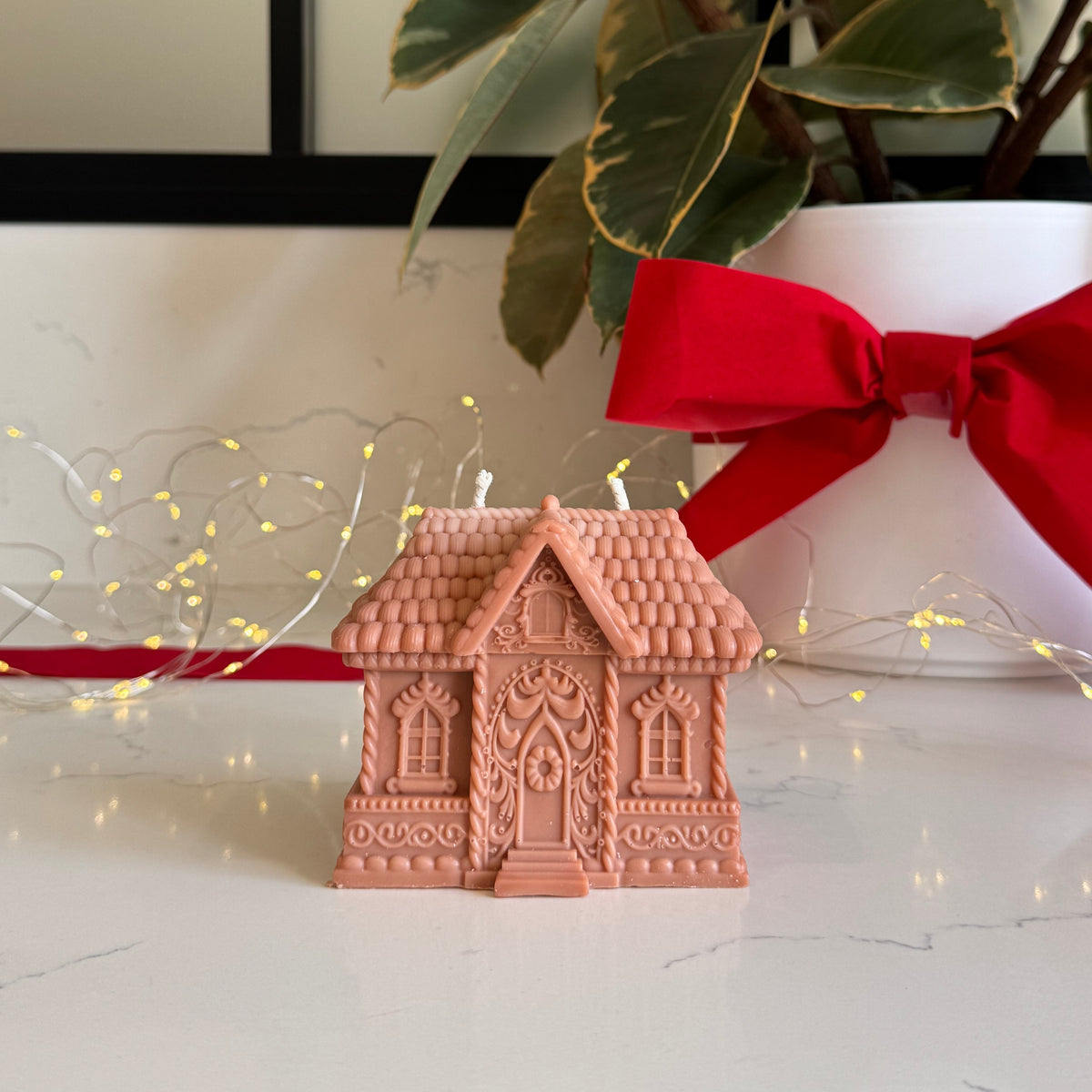 A small light brown gingerbread house-shaped candle sits on a white marble surface, styled with a red ribbon bow, a potted plant, and delicate fairy lights in the background.