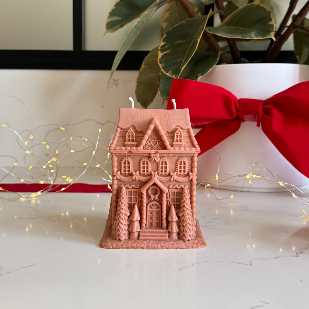 A large gingerbread house-shaped candle in a light brown color sits on a white marble surface, styled with a red ribbon, a potted plant, and warm string lights in the background.