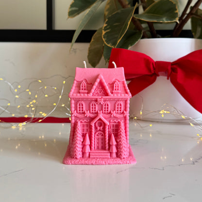 A pink gingerbread house-shaped candle sits on a white surface in front of a white planter with green variegated leaves and a large red bow. Warm string lights are visible in the background.