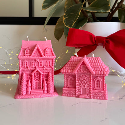 Two pink gingerbread house-shaped candles displayed on a white marble surface, with a white pot, green leaves, and a red ribbon in the background.