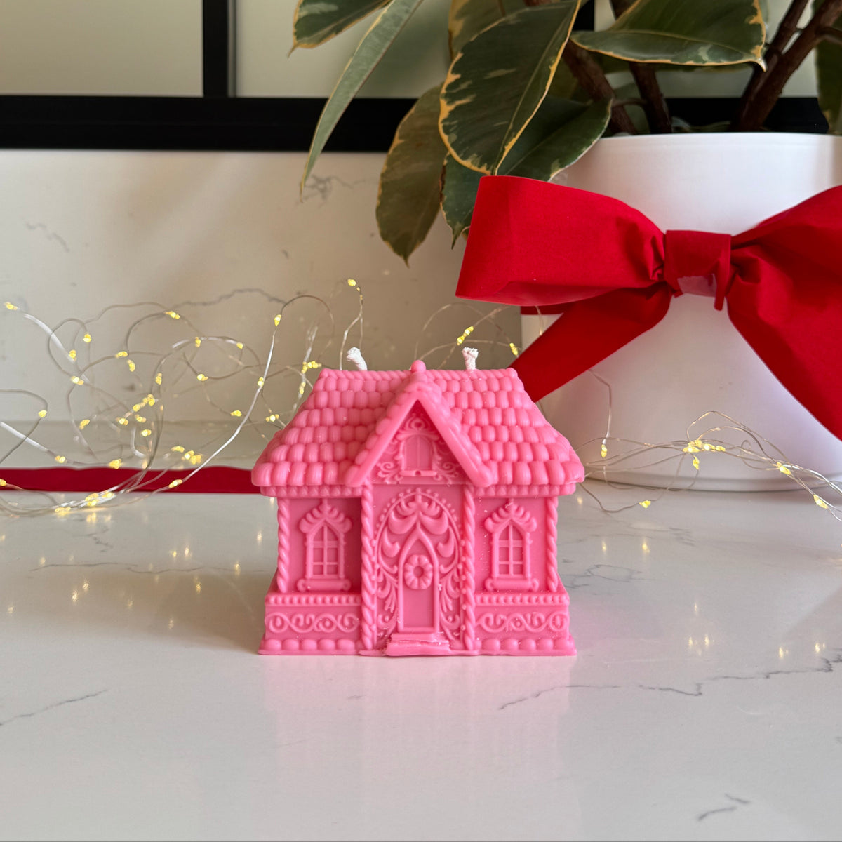 A pink gingerbread house-shaped candle sits on a glossy white marble surface, styled with a red ribbon and bow, green leaves, and warm string lights in the background.