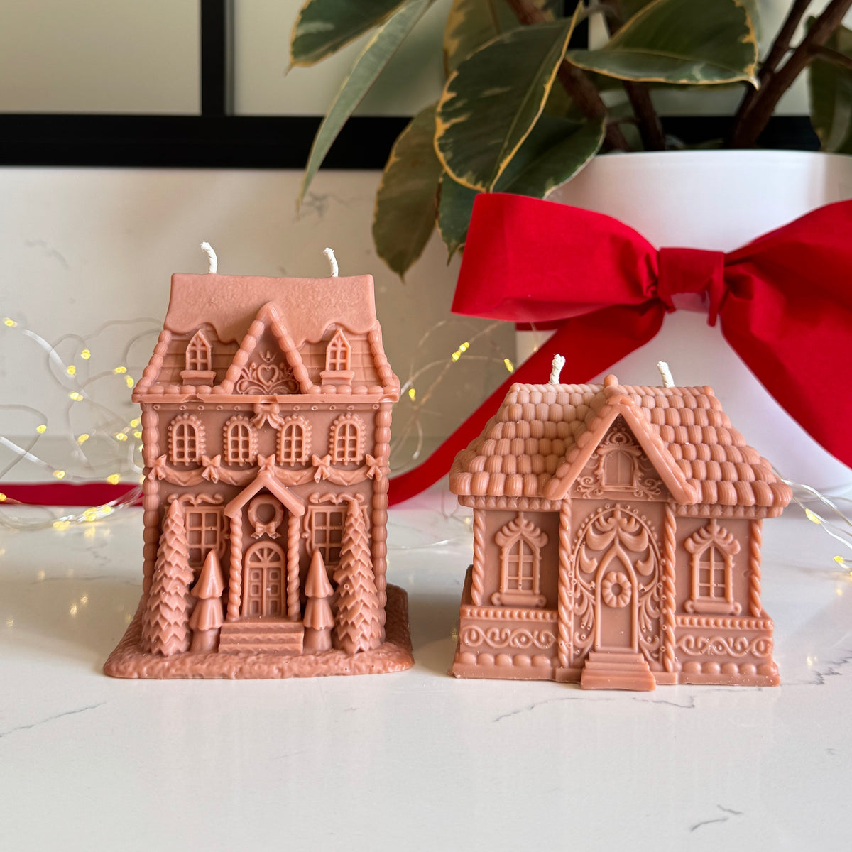 Two decorative gingerbread house-shaped candles in a light brown color, displayed on a white marble surface with a white pot, green leaves, and a red ribbon in the background.