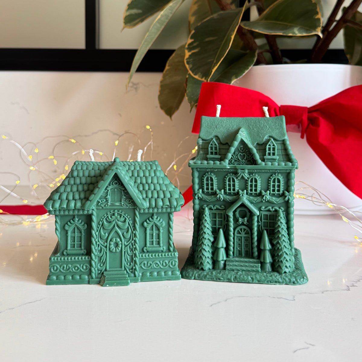 Two green gingerbread house-shaped candles displayed on a white surface, with a potted plant and red ribbon in the background.