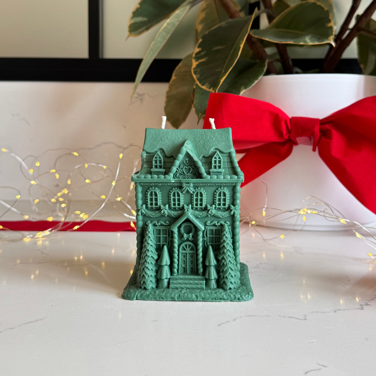A green gingerbread house-shaped candle with two white wicks sits on a white marble surface, styled with a red ribbon, a potted plant, and warm fairy lights in the background.