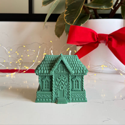 A green gingerbread house-shaped candle sits on a white surface in front of a white planter with a large red bow and green variegated leaves. Warm string lights are visible in the background.