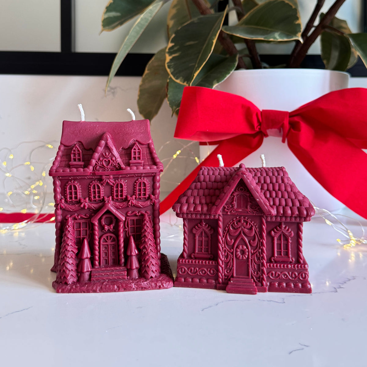Two red gingerbread house-shaped candles on a white surface, with one larger ornate house candle and one smaller house candle tied with a red ribbon. A potted plant and string lights are blurred in the background.