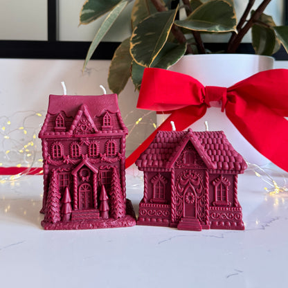 Two red gingerbread house-shaped candles on a white surface, with one larger ornate house candle and one smaller house candle tied with a red ribbon. A potted plant and string lights are blurred in the background.