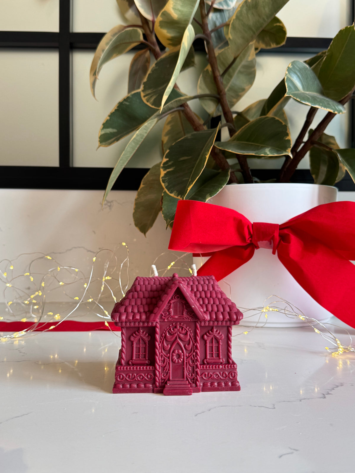A small red gingerbread house-shaped candle sits on a white marble surface, styled with a red ribbon, fairy lights, and a potted plant in the background.