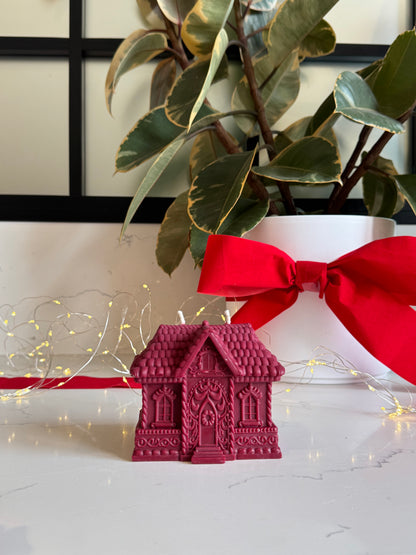 A small red gingerbread house-shaped candle sits on a white marble surface, styled with a red ribbon, fairy lights, and a potted plant in the background.