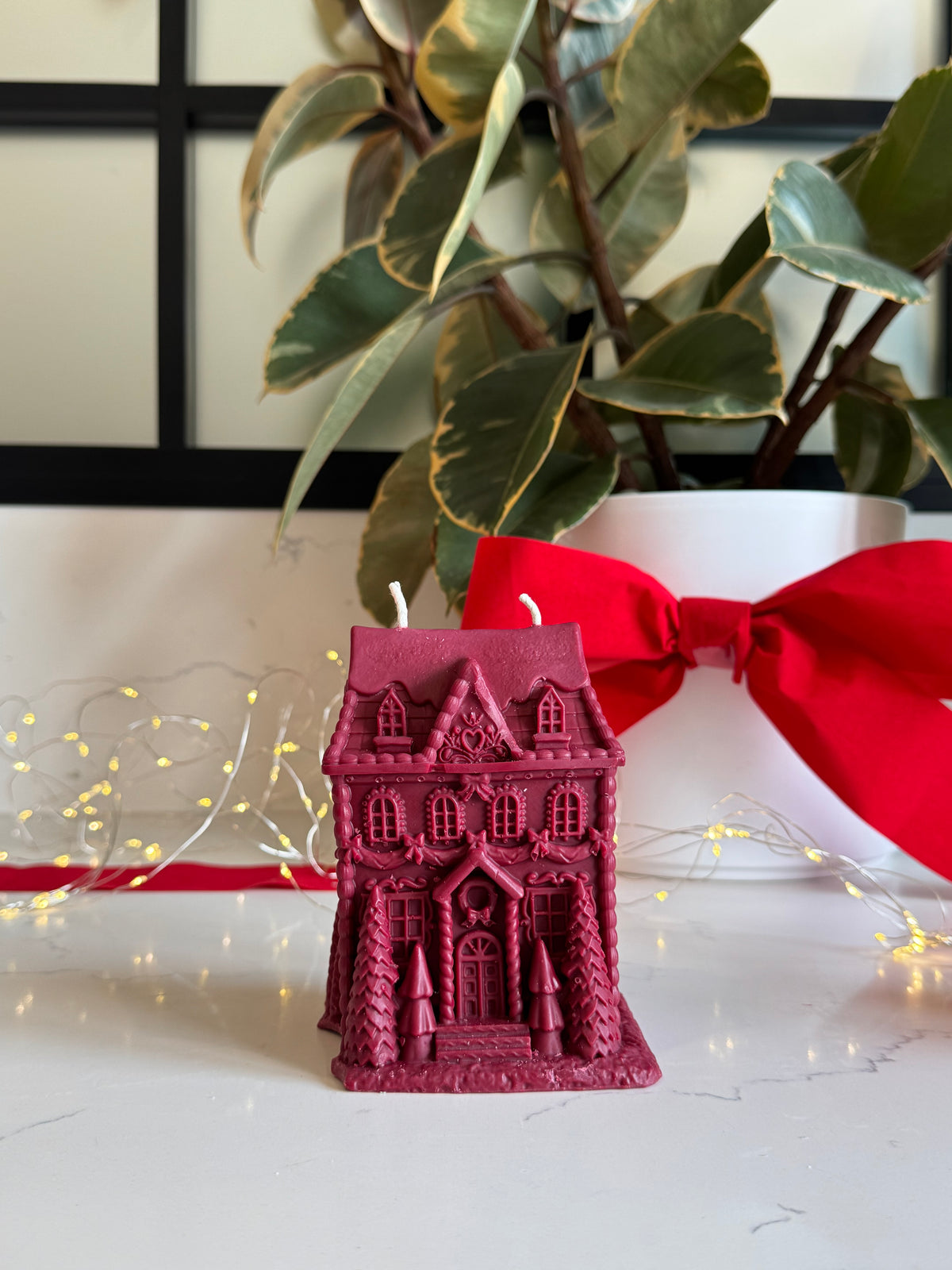 A small magenta gingerbread house-shaped candle with two wicks sits on a white surface in front of a white planter with green variegated leaves and a large red ribbon. Warm string lights are visible in the background.