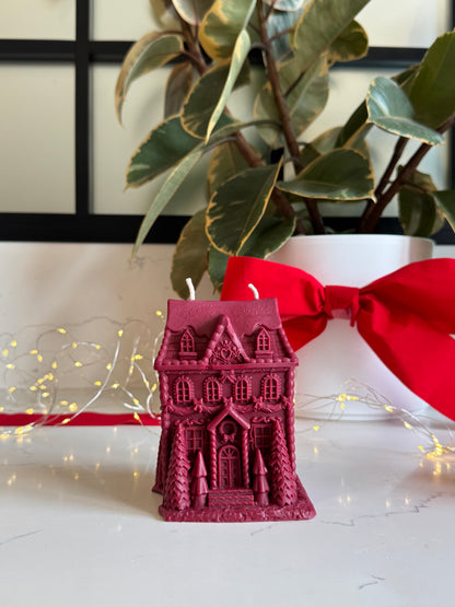 A small magenta gingerbread house-shaped candle with two wicks sits on a white surface in front of a white planter with green variegated leaves and a large red ribbon. Warm string lights are visible in the background.