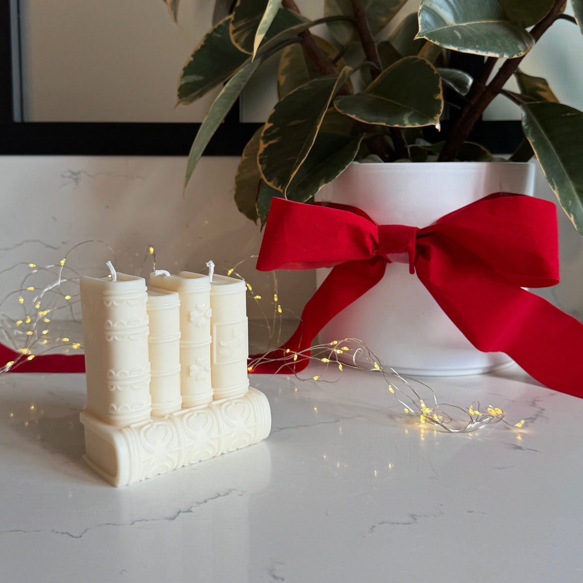 Book Shaped decorative candle with 5 books arranged on a white marble surface next to a white planter with a large green plant and a red ribbon, decorated with warm fairy lights.