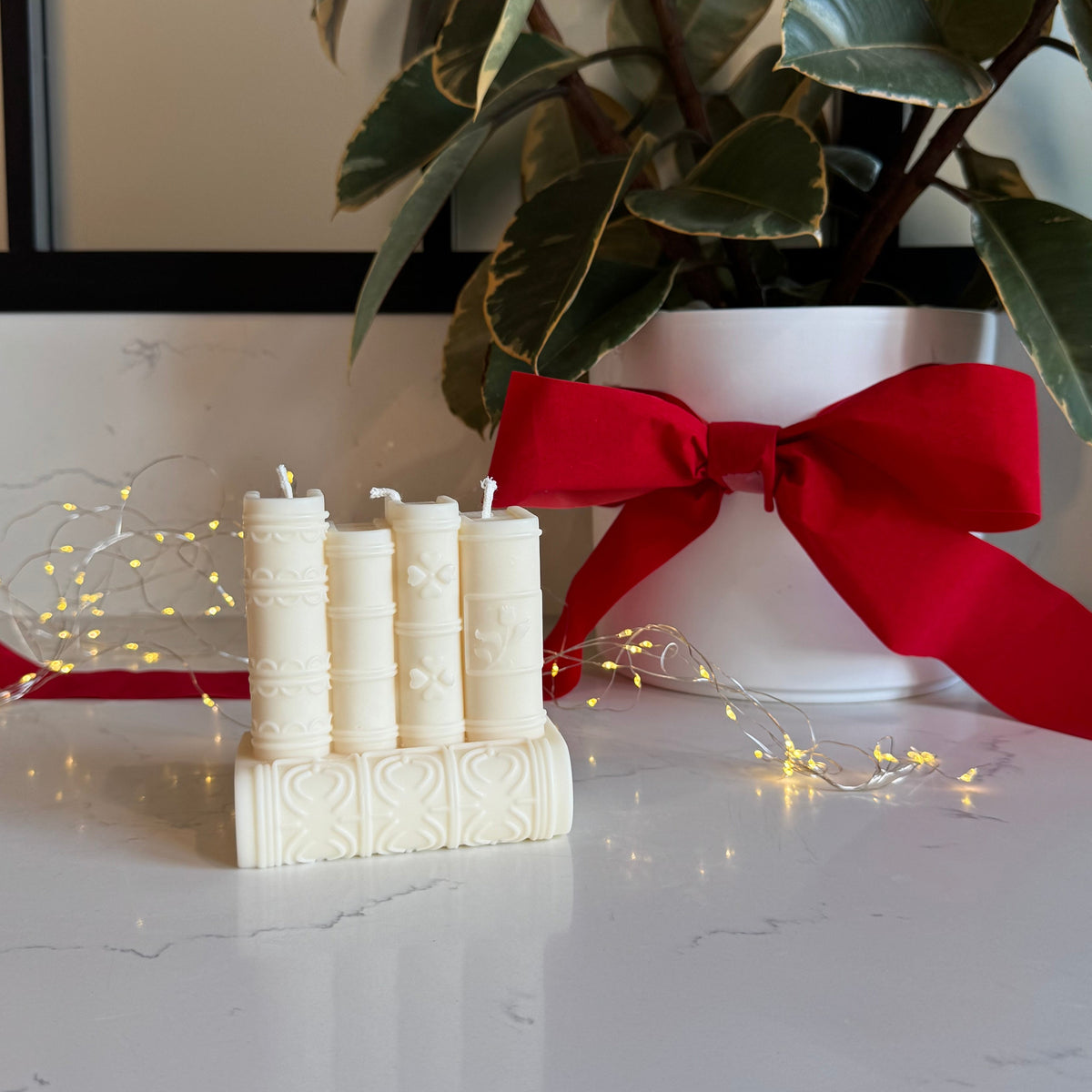A stack of five books shaped candle arranged on a white marble surface, styled with a red ribbon bow on a white planter with green variegated leaves and warm fairy lights in the background.
