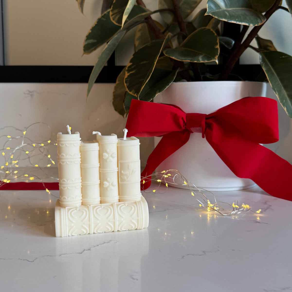 A set of five cream-colored book shaped decorative candles arranged on a white marble surface, with a red ribbon bow and a potted variegated plant in the background.
