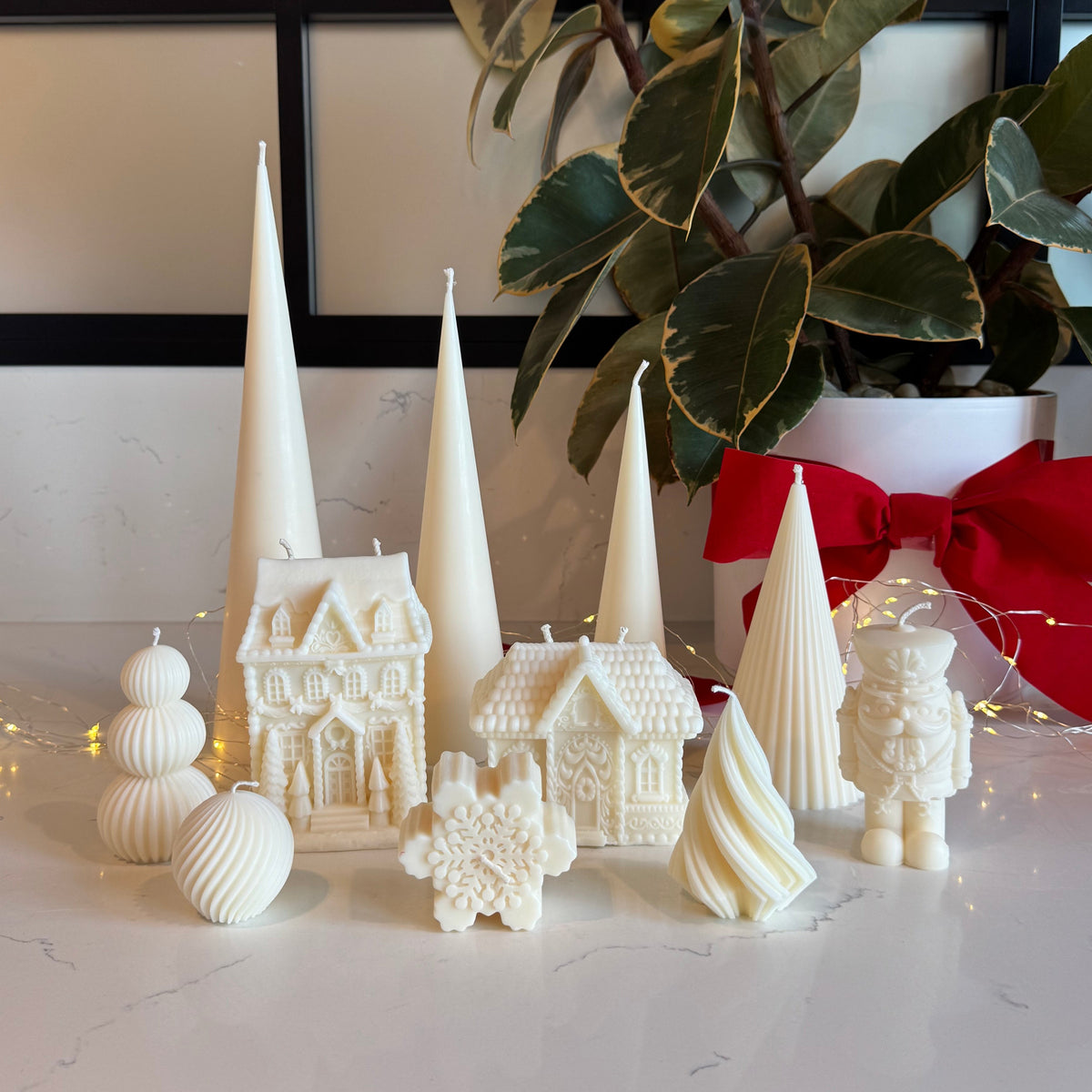 A collection of white holiday-themed candles shaped like houses, trees, snowballs, and a nutcracker, arranged on a white marble countertop next to a potted magnolia plant with a red bow and warm string lights.