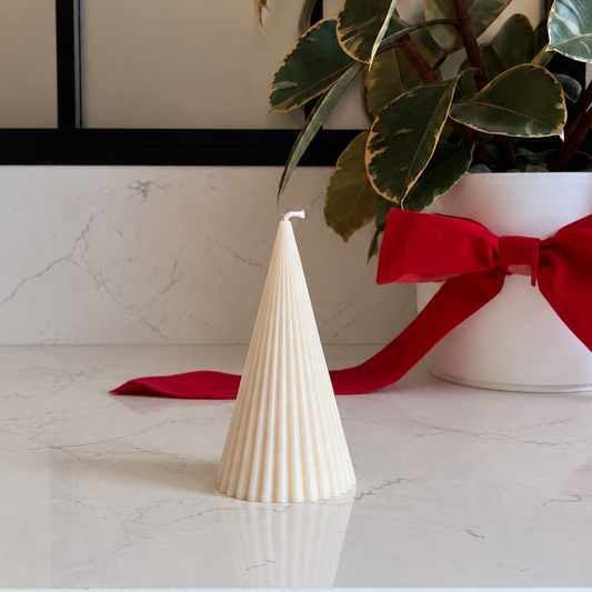 A cream-colored, ribbed Christmas tree-shaped candle sits on a white marble countertop next to a potted rubber plant with a red ribbon bow.